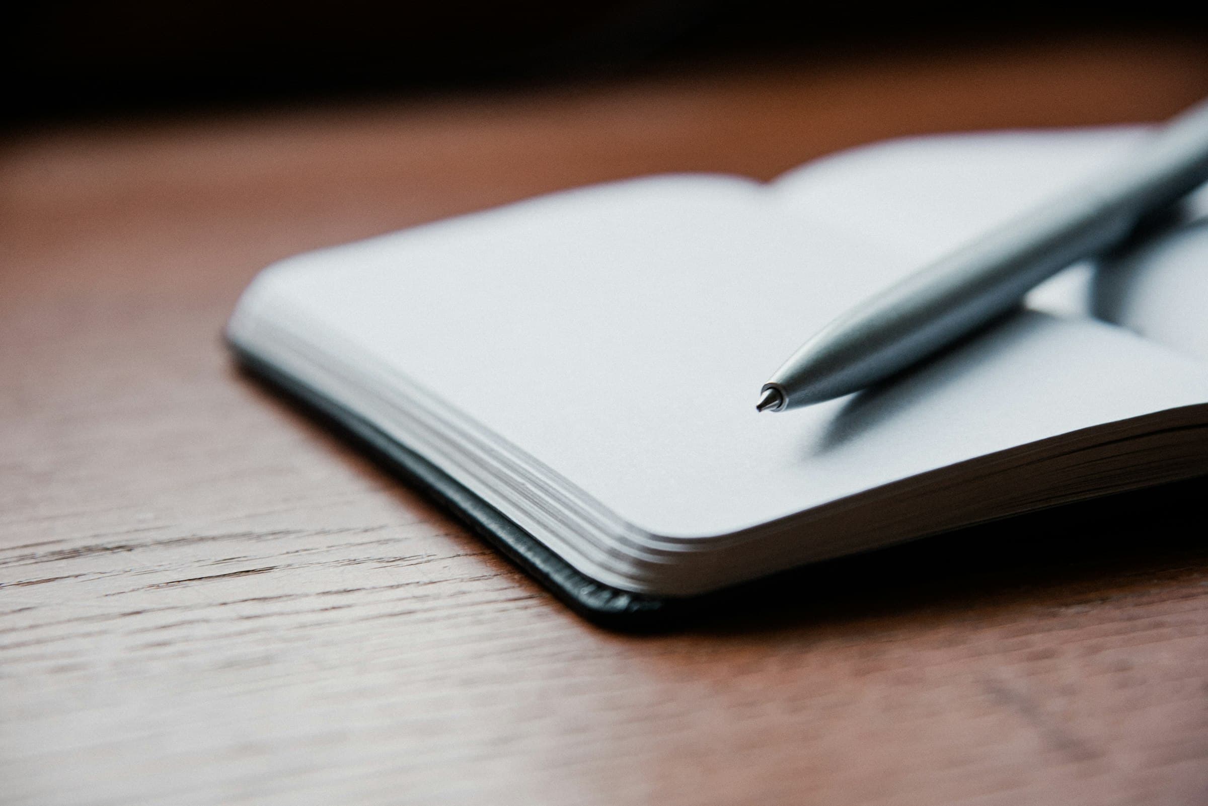 Libreta abierta con las hojas en blanco y una pluma plateada, sobre una mesa de madera